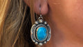 A close-up of a person wearing a silver-toned Southwestern-style dangle earring with a bright turquoise blue oval stone, shown hanging from the earlobe with part of a matching necklace visible below.