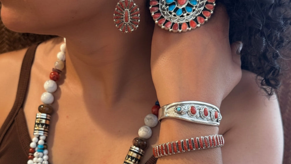 A woman in a brown tank top models a set of handmade jewelry, featuring a large round turquoise and red beaded earring, a multi-strand white bead necklace, and a silver cuff bracelet with turquoise and red accents.